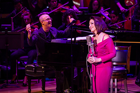 A man smiling in the background while he plays the piano and a woman in a pink dress smiles behind a microphone